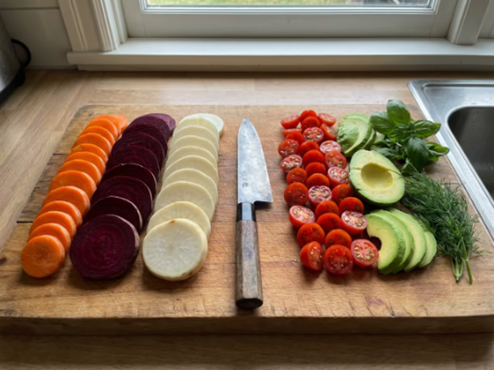 Cortar verduras: ¿Cómo hacerlo rápido y fácil? 8 Tabla de cortar con verduras cortadas en rodajas: zanahoria, remolacha, nabo, tomates cherry, aguacate y hierbas frescas junto a cuchillo de chef